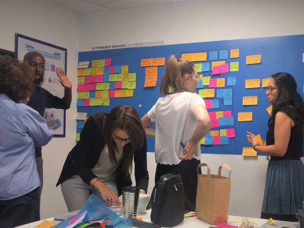 Photo of 5 people talking in front of a colourful 'agile' wall with post-its on