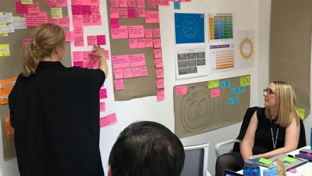 three members of a team looking at an agile wall filled with colourful posters and post-it notes