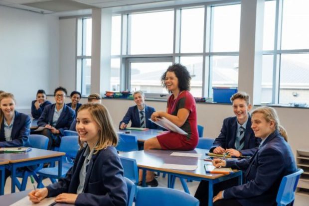 Teacher sat amongst classroom of (Key Stage 3 or 4 it's not determined) pupils all looking towards the front of the room. Everyone is looking very engaged and happy.