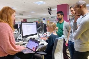 A photo of 5 members of a team stood looking at a screen. One member of the team also has their laptop which shows a Trello Board. 