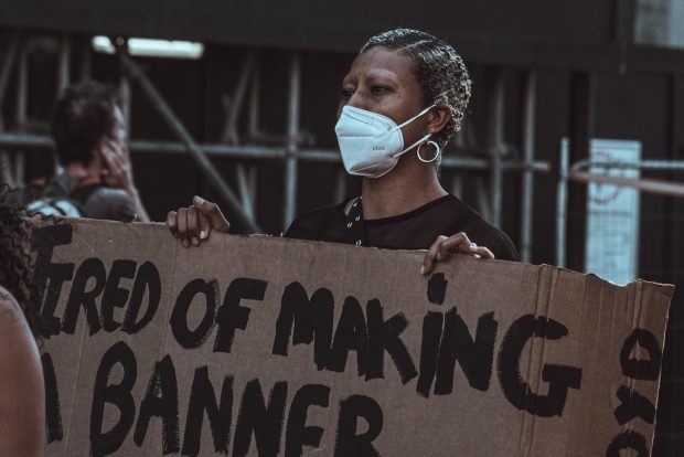 Image of a black woman wearing a face mask and holding a sign which reads 'Tired of making a banner'