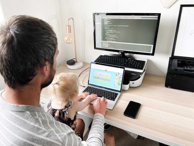 A behind the head shot of Steven looking at a laptop screen of code and his daughter, Kitty, sitting on his lap looking at a smaller screen of Peppa pig