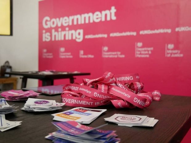 The focus of the image are some lanyards and sticker on a table. They're in front of a pink billboard that reads 'Government is hiring'