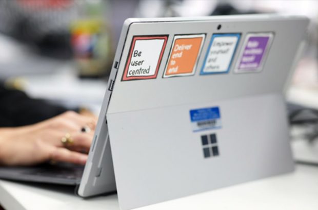 Back of a laptop with 4 stickers on. Only the first two stickers are in focus, reading 'be user centred' and 'deliver end to end'. A white hand is typing on the laptop