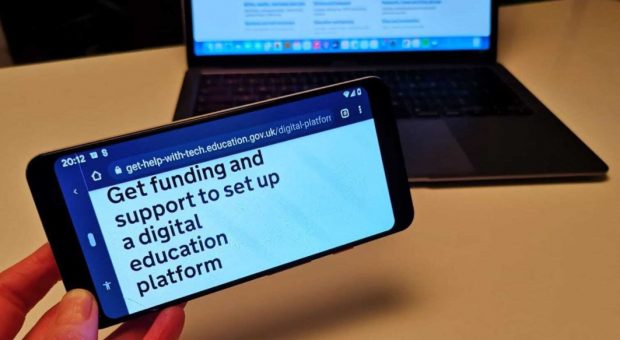 GOV.UK web page on a mobile phone screen in front of a laptop. Mobile phone reads 'Get funding and support to set up a digital education platform'. 