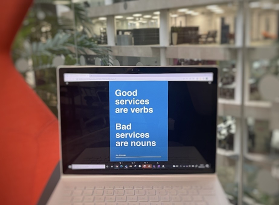 white text on a blue computer screen reading 'good services are verbs' and 'bad services are nouns'. image of an office atrium blurred in the background