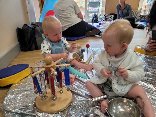 Two babies at a Family Hub, playing with different kinds of toys, sitting on a matt.