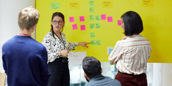 members of a team stood around looking at a board with colourful posting notes. one member is pointing at one in particular